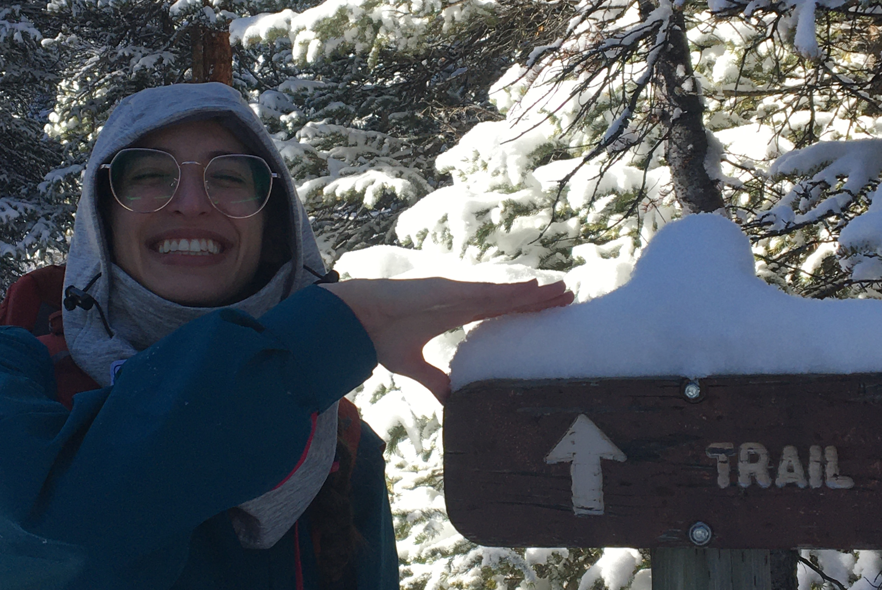 juniper wearing a gray balaclava and grinning at the camera while showing how much snow has accumulated on a trail sign.